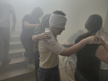 Szkolenie Bezpieczna Ewakuacja organizowane przez BON UWM, na zdjęciu zadymiona klatka schodowa, na której osoby idące gęsiego trzymające się jeden drugiego schodzą z zadymionej klatki schodowej, w pierwszym kadrze jedna z osób ma zawiązane przepaską oczy, w drugim kadrze widoczna osoba na wózku inwalidzkim znoszona ze schodów klatki schodowej przez inne osoby.
