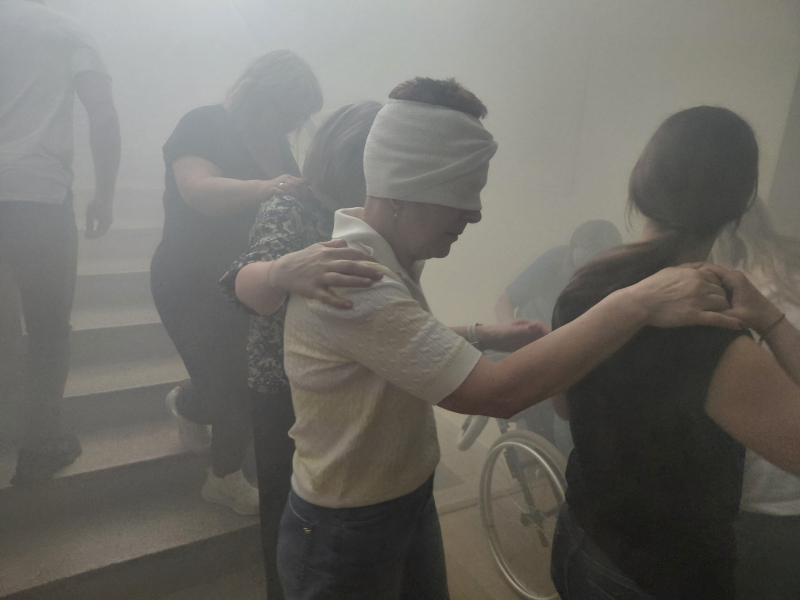 Szkolenie Bezpieczna Ewakuacja organizowane przez BON UWM, na zdjęciu zadymiona klatka schodowa, na której osoby idące gęsiego trzymające się jeden drugiego schodzą z zadymionej klatki schodowej, w pierwszym kadrze jedna z osób ma zawiązane przepaską oczy, w drugim kadrze widoczna osoba na wózku inwalidzkim znoszona ze schodów klatki schodowej przez inne osoby.